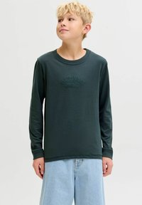 Tamna zelena majica dugih rukava s okruglim izrezom i reljefnim natpisom "ORIGINALS" na prednjem dijelu, u kombinaciji sa svijetlim trapericama.