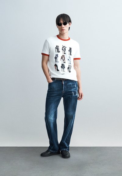 Άνδρας με γυαλιά ηλίου, λευκό μπλουζάκι με πολύχρωμες εκτυπώσεις ψηλοτάκουνων παπουτσιών, μπλε τζιν και μαύρα παπούτσια, στέκεται σε ένα απλό φωτεινό τοίχο.