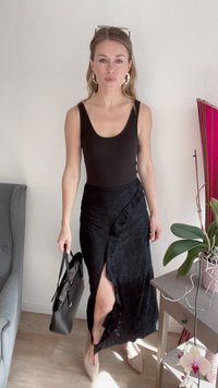 Body preto sem mangas combinado com uma saia envolvente texturizada preta com uma fenda até a coxa. Acessório com uma bolsa preta; usando sapatos nude.
