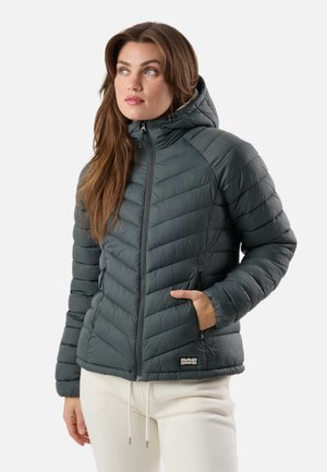 Frau mit langem Haar, die eine dunkelgraue gesteppte Kapuzenjacke mit Reißverschlusstaschen und cremefarbene Hose trägt, schaut leicht nach oben und zur Seite.