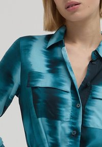 Camicia tie-dye turchese e nera, maniche lunghe, design con bottoni, due tasche sul petto, texture simile alla satin, e colletto a punta.