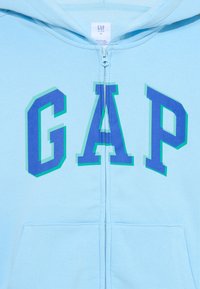 Lyseblå hettegenser med glidelås, som har en fet "GAP"-logo i dyp blått med grønne detaljer. Laget av myk bomullsblanding, inkluderer en frontlomme.