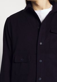 Navy button-up shirt in a soft, textured fabric. Features two chest pockets and embroidered branding on the front side.