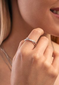 Silberner, gedrehter Ring an einem Finger. Glatte, glänzende Textur mit geflochtenem Design. Haut ist sichtbar und reflektiert Licht in sanften Tönen.