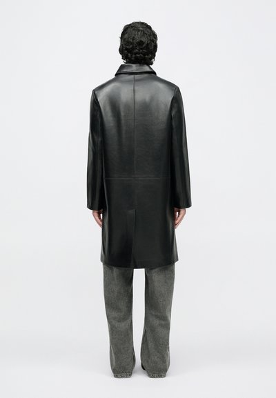 Kiharaliste juustega mees, kes kannab musta nahast pikka mantlit ja hallid laia säärega pükse, seisab seljaga vaataja poole valgel taustal.