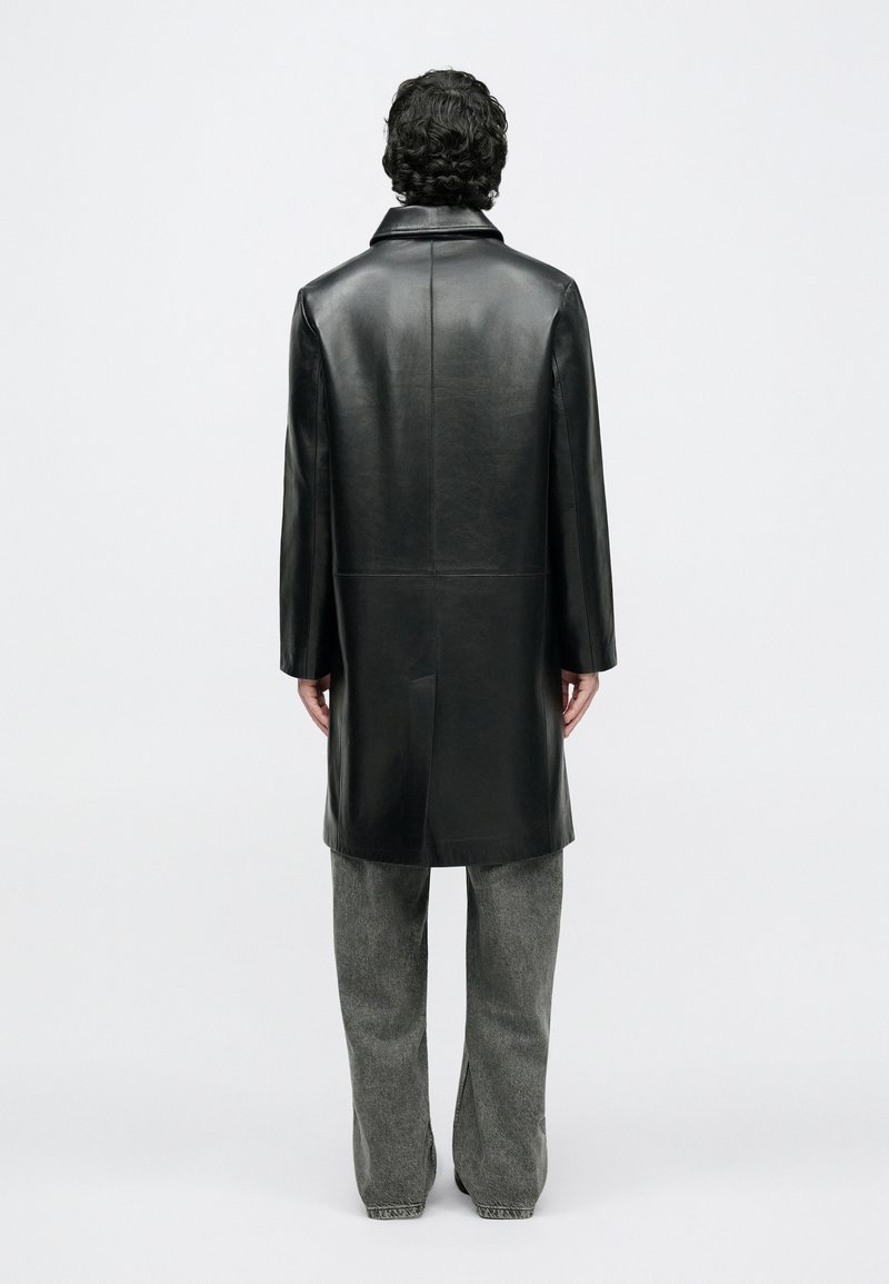 Homme aux cheveux bouclés portant un manteau long en cuir noir et un jean gris à jambes larges, debout de dos sur un fond blanc uni.