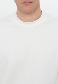 Fehér kerek nyakú pulóver, melynek mellén texturált "ARMANI EXCHANGE" logó található. Puha pamut anyagból készült, hétköznapi viseletre tervezve.
