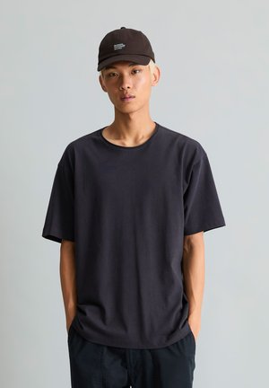 Jonge man met lichtblond haar, die een zwarte pet, zwart T-shirt en zwarte broek draagt, staand tegen een effen grijze achtergrond.