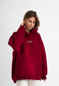 Felpa oversize color bordeaux con tasca frontale, con testo ricamato bianco "ESSERE UNICO ESSERE SE STESSO" sul petto. Tessuto morbido.