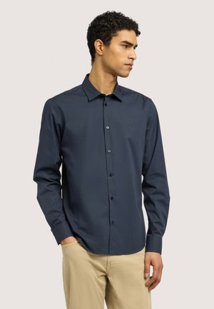 Nuori mies, jolla on tumma laivastonsininen nappipaita ja beige housut, katsoo oikealle yksinkertaista vaaleaa taustaa vasten.