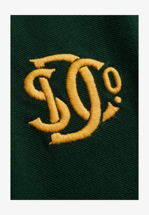 Gult stiliseret "S" og "O²" broderi på mørkegrøn tekstureret stofbaggrund, nærbillede.