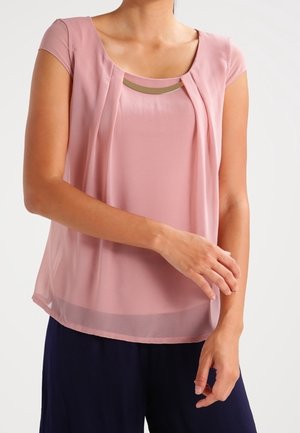 Femme portant un chemisier sans manches rose poussiéreux avec un détail doré au niveau du col, associé à un pantalon bleu foncé, sur un fond uni.