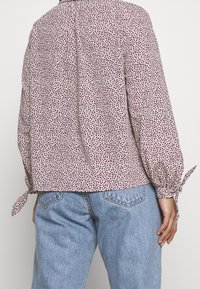 Långärmad blus i lätt tyg med en krämfärgad bakgrund och spridda vinröda prickar. Har knytärmar och en avslappnad passform.