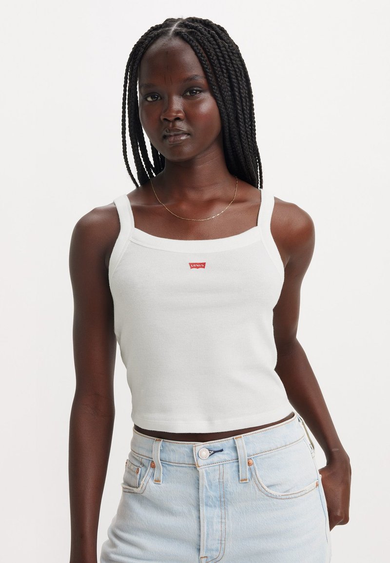 Bílý ribbed crop top s čtvercovým výstřihem a tenkými ramínky, na přední straně s malým červeným logem Levi's. Nesený s světle modrými denimovými šortkami.