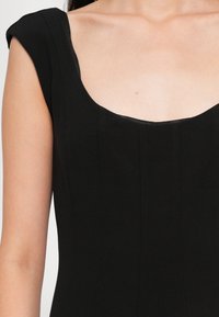 Robe noire avec un décolleté carré et des manches courtes. Le tissu est lisse avec des détails de coutures verticales et une silhouette ajustée.