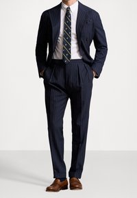 Marineblauw krijtstreeppak met een dubbelgeknoopt colbert en geplooide broek, gecombineerd met een patroonstropdas, wit overhemd en bruine loafers.