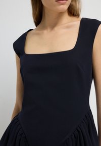 Femme portant une robe sans manches de couleur bleu marine foncé avec un décolleté carré et un détail de tissu froncé à la taille, vue des épaules aux hanches.