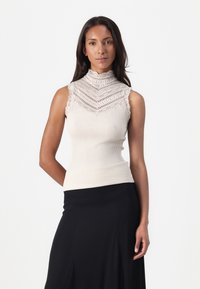 Ribbad krämfärgad ärmlös topp med hög krage, dekorerad med intrikata spetsdetaljer och en figurnära design, i kombination med en svart flödande kjol.