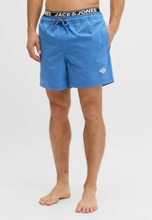 Uomo che indossa pantaloncini da bagno blu fino a metà coscia con coulisse, tasche laterali e cintura con marchio Jack & Jones, in piedi a piedi nudi su sfondo bianco.