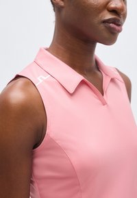 Κοντινό πλάνο ενός ατόμου που φοράει ροζ τοπ χωρίς μανίκια με γιακά, εμφανίζοντας τον ώμο, τον λαιμό και μερικό πρόσωπο μπροστά σε απλό φόντο.