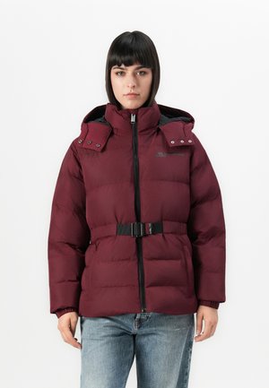 Vrouw in een maroon pufferjack met een capuchon en riem, staand met ontspannen armen, tegen een eenvoudige witte achtergrond.