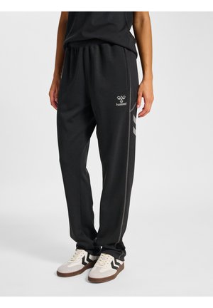 Person iført sorte Hummel træningsbukser med sidestriber og beige sneakers med sorte chevronmønstre.