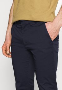 Marinblå byxor i bomull med en slät yta, sidofickor och en figurnära design. Bärs med en beige topp.