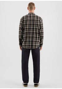 Camicia a quadri nera, marrone e bianca, con colletto button-down e maniche lunghe, abbinata a jeans scuri e scarpe casual.