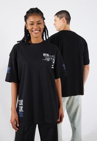 Black oversized t-shirt made of cotton, featuring graphic prints in white and blue on the front and left sleeve. Casual fit with short sleeves.