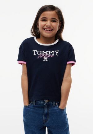 Ragazza sorridente con capelli castani di media lunghezza indossa una maglietta blu navy Tommy Hilfiger con bordi bianchi e rosa, mani in tasca, in piedi davanti a uno sfondo bianco.
