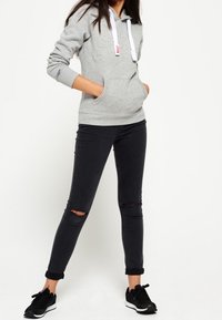 Felpa grigia con coulisse bianche e tasca frontale, abbinata a jeans skinny neri strappati e scarpe da ginnastica nere. Outfit semplice e casual.