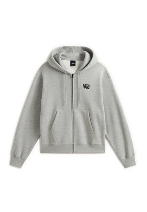 Jeune homme aux cheveux bruns et raides porte un sweat à capuche zippé Vans gris clair et un pantalon beige, debout devant un fond uni.