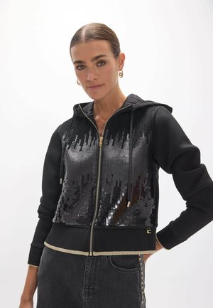 Mujer con sudadera negra con cremallera y lentejuelas negras brillantes en la parte delantera y vaqueros oscuros con tachuelas laterales, de pie sobre un fondo blanco.
