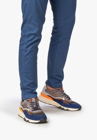 Scarpe sportive grigie e blu con accenti arancioni, caratterizzate da una tomaia in rete e camoscio, una suola bianca spessa e lacci decorati.