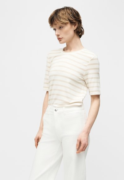 Γυναίκα που φοράει μπεζ και λευκό κοντομάνικο πουκάμισο με οριζόντιες ρίγες και λευκό ψηλόμεσο παντελόνι, στέκεται μπροστά σε απλό ανοιχτόφωτο φόντο.