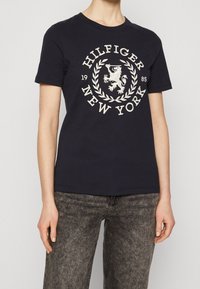 Marineblå bomulds T-shirt med et hvidt broderet logo med en løve og laurbær, rund halsudskæring og korte ærmer. Bæres med grå jeans.