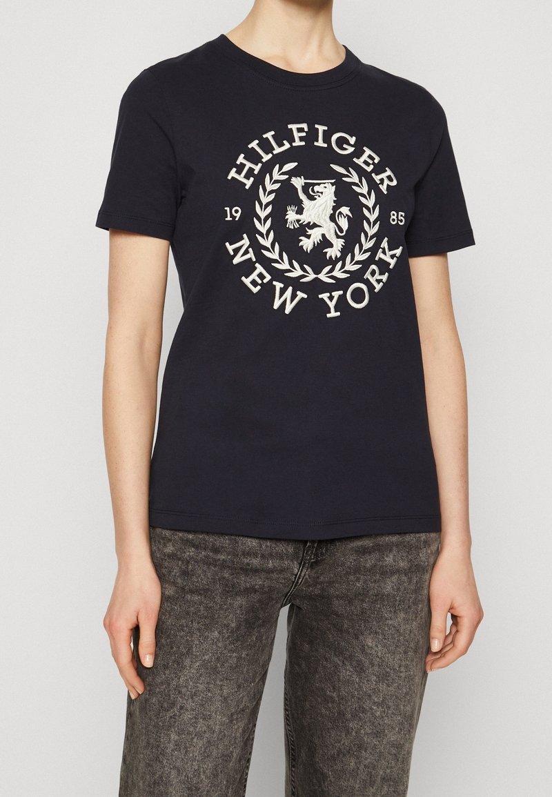 Marineblå bomulds T-shirt med et hvidt broderet logo med en løve og laurbær, rund halsudskæring og korte ærmer. Bæres med grå jeans.