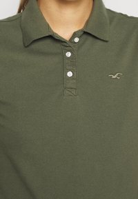 Olivgrön polo skjorta i texturerad bomull, med en treknäppningsslå och en liten broderad logotyp på bröstet.