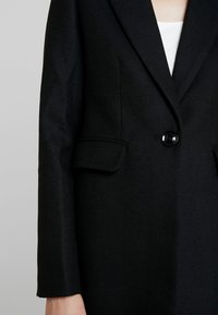 Černý sako na míru s jedním knoflíkem a klopovým kapsou na levé straně, nošený přes bílý top.