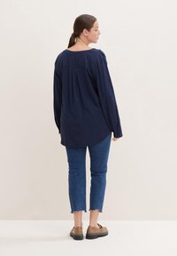 Blusa de manga larga azul marino con recogidos en los hombros, de tela suave, con dobladillo curvado. Llevada con pantalones vaqueros azules cortos y zapatos marrones con suelas negras.