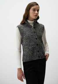 Gilet grigio in maglia con colletto, bottoni frontali e due tasche, abbinato a una maglietta bianca a maniche lunghe e pantaloni neri.