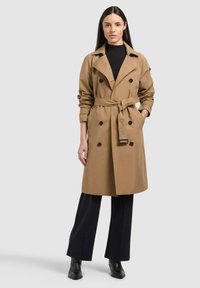 Donna con capelli lunghi e scuri che indossa un trench beige con cintura sopra un dolcevita nero, pantaloni neri e stivaletti neri, in piedi di fronte.