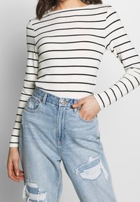 Långärmad, krämfärgad topp med svarta horisontella ränder, parat med ljusblå slitna jeans med fransade kanter och fickor.