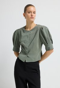 Frau mit nach hinten gekämmtem Haar trägt eine olivgrüne, geknöpfte Bluse mit Puffärmeln und schwarze, hochtaillierte Hosen vor einem weißen Hintergrund.