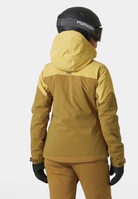 Geel en bruine skijas met een capuchon, lange mouwen en zwarte handschoenen. Bevat contrasterende boven- en onderdelen met een logo op de achterkant.
