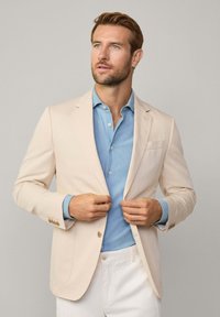 Blazer beige clair avec deux boutons, revers crantés et poche plaquée. Associé à une chemise bleu clair et un pantalon blanc, créant un look superposé.