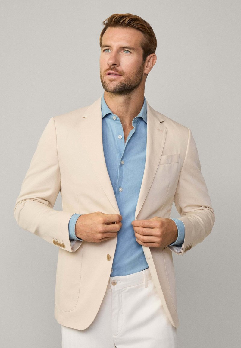 Lichtbeige blazer met twee knopen, notch revers en opgestikte zak. Gepaard met een lichtblauw overhemd en witte broek, wat een gelaagde look creëert.