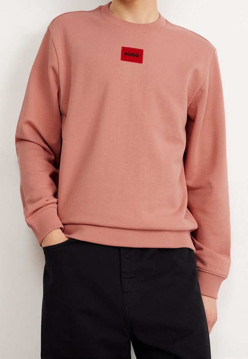 Personne portant un sweat-shirt rose pâle avec un patch logo rouge "HUGO" sur la poitrine et un pantalon noir, une main dans la poche.