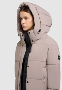 Puffige beige Jacke mit großer Kapuze, Druckknöpfen und Reißverschluss. Verfügt über Seitentaschen und ein Logopatch am Ärmel, glatte Textur.