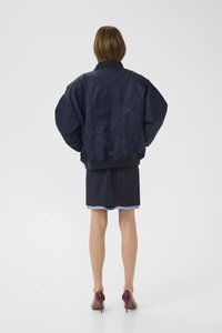 Navy bomber jacket with a textured finish, oversized shape, and ribbed cuffs. Back features a subtle logo detail, worn over a pinstriped skirt.